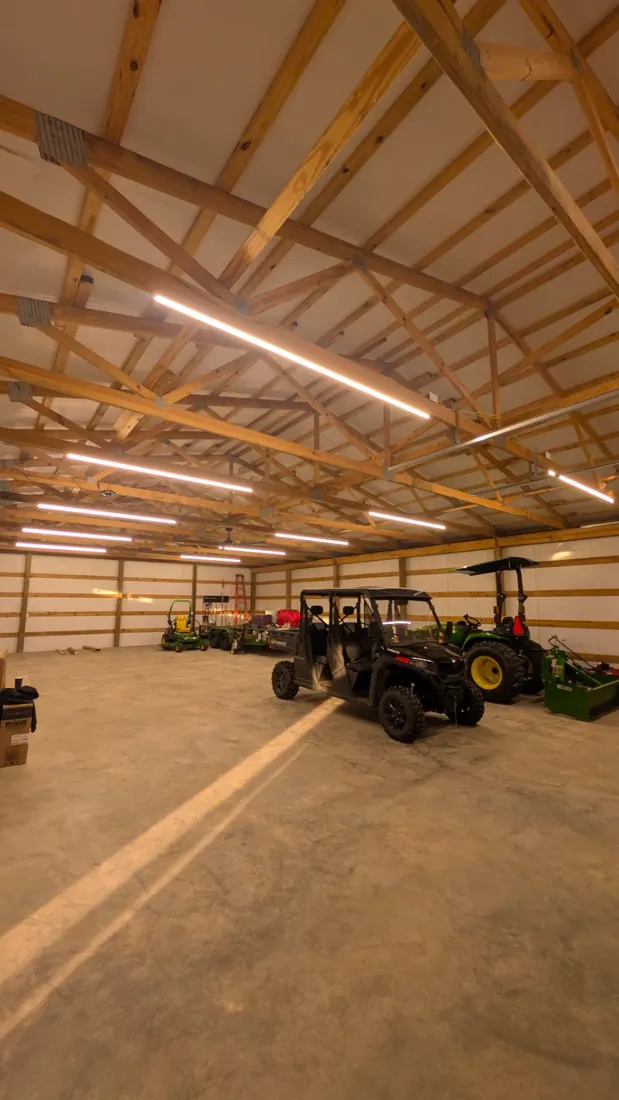 Long LED strip lights running along the wood trusses of a pole barn with UTVs parked on the concrete floor